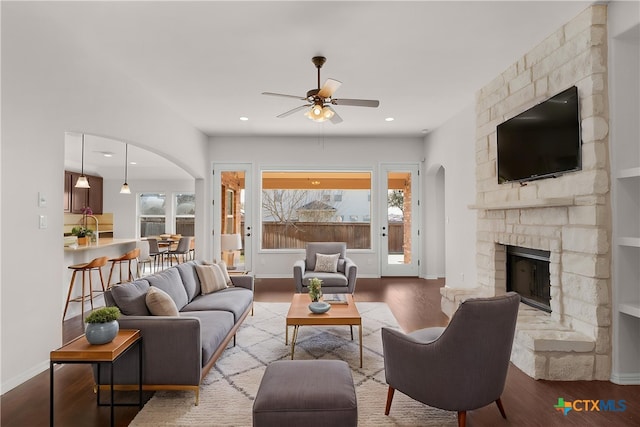 living area featuring arched walkways, wood finished floors, a ceiling fan, and a stone fireplace
