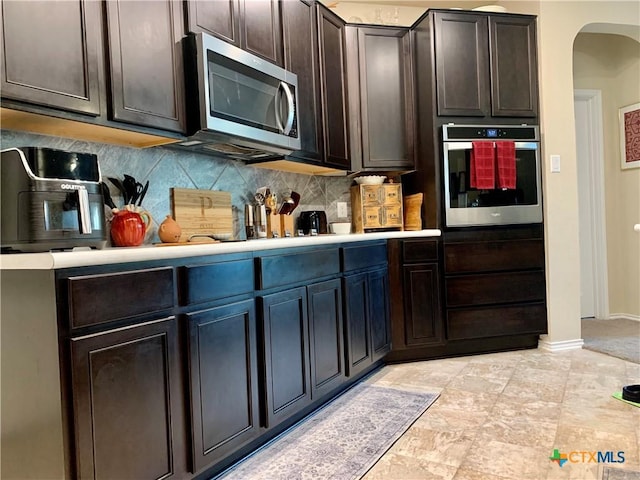 kitchen with arched walkways, stainless steel appliances, light countertops, backsplash, and dark brown cabinets