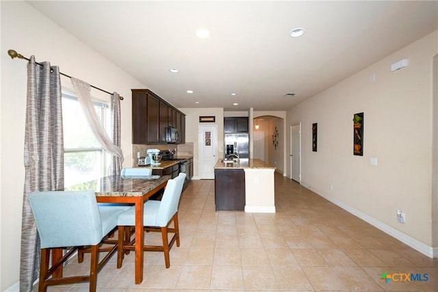 kitchen featuring appliances with stainless steel finishes, light tile patterned floors, dark brown cabinetry, arched walkways, and a kitchen island with sink