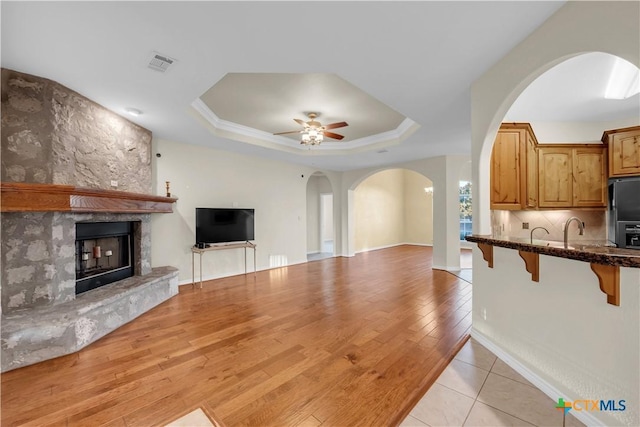 unfurnished living room featuring a raised ceiling, ceiling fan, a stone fireplace, and light wood-type flooring