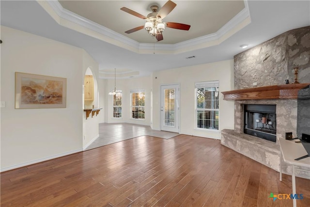 unfurnished living room featuring a tray ceiling, a stone fireplace, wood-type flooring, and ceiling fan with notable chandelier