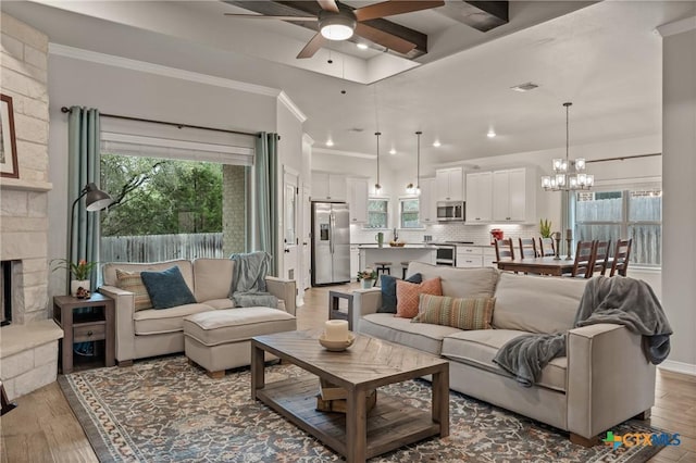 living room with visible vents, light wood-style flooring, ornamental molding, a stone fireplace, and ceiling fan with notable chandelier