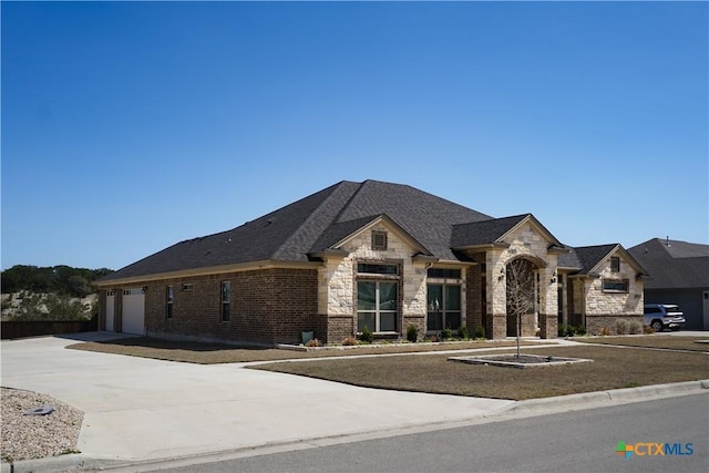 french provincial home with brick siding, an attached garage, roof with shingles, stone siding, and driveway