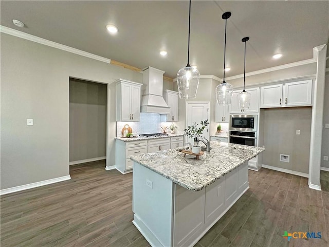 kitchen with custom exhaust hood, white cabinetry, stainless steel appliances, light stone countertops, and a kitchen island with sink