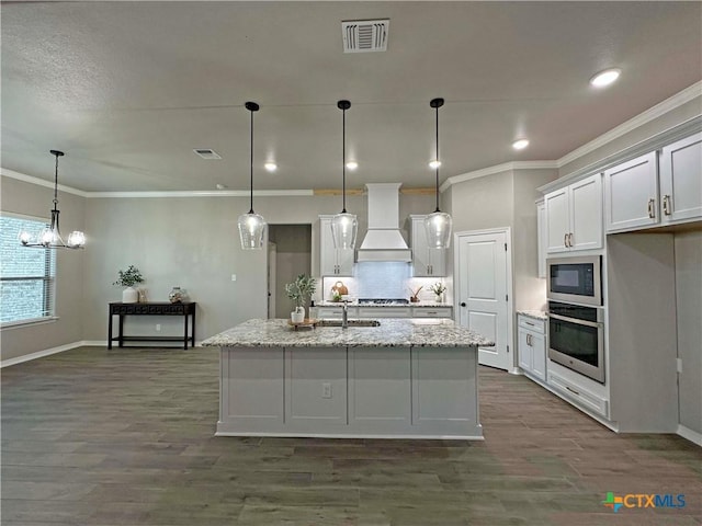 kitchen featuring appliances with stainless steel finishes, custom range hood, decorative light fixtures, light stone countertops, and a kitchen island with sink