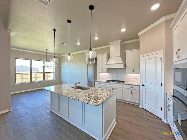 kitchen featuring hanging light fixtures, light stone counters, premium range hood, a center island with sink, and white cabinets