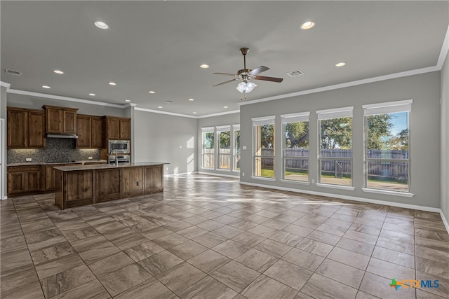 kitchen with stainless steel microwave, decorative backsplash, a kitchen island with sink, ceiling fan, and crown molding