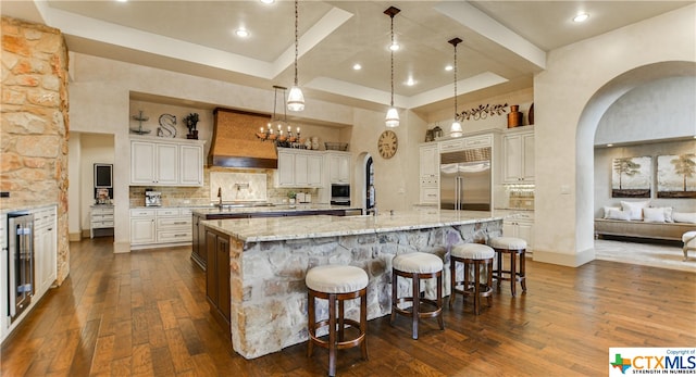 kitchen featuring dark wood-type flooring, stainless steel built in refrigerator, decorative light fixtures, and a large island with sink