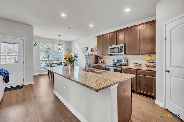 kitchen featuring a kitchen island with sink, a sink, tasteful backsplash, appliances with stainless steel finishes, and light stone countertops