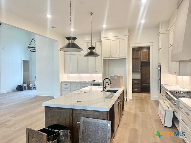 kitchen featuring a sink, a kitchen island with sink, light wood-style floors, white cabinetry, and custom range hood