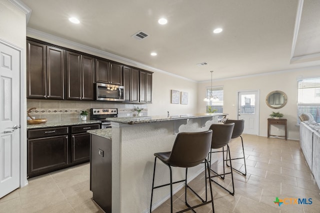 kitchen featuring dark brown cabinetry, appliances with stainless steel finishes, light stone countertops, decorative light fixtures, and an island with sink