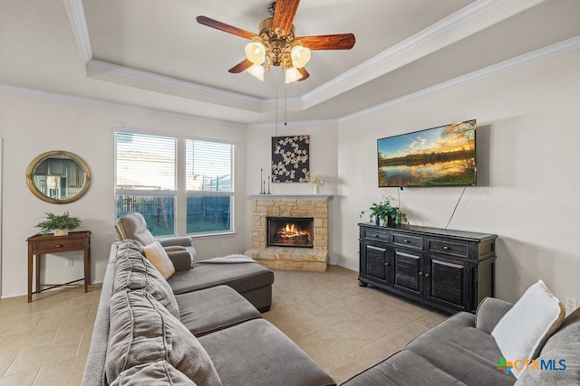 tiled living room featuring a stone fireplace, a raised ceiling, ceiling fan, and crown molding