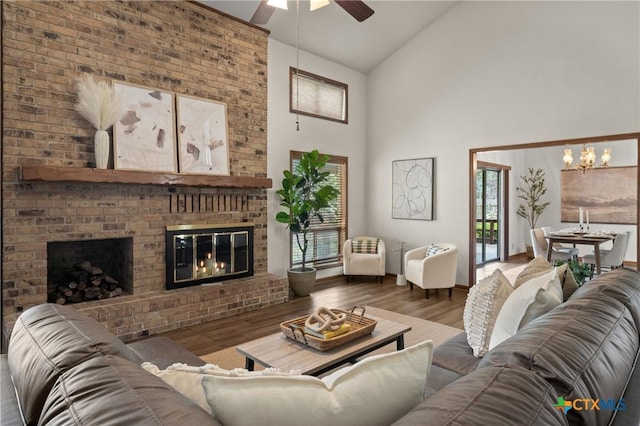 living room with a brick fireplace, high vaulted ceiling, wood finished floors, and ceiling fan with notable chandelier