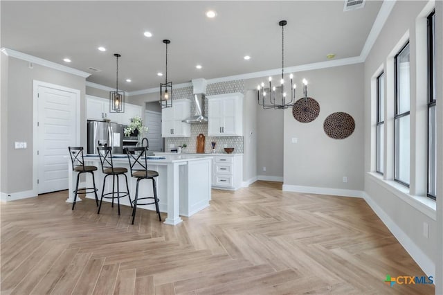 kitchen featuring hanging light fixtures, stainless steel fridge, wall chimney exhaust hood, a kitchen island with sink, and white cabinets