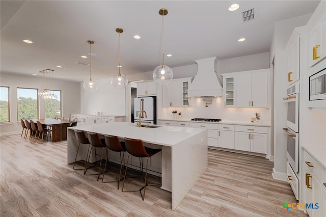 kitchen featuring custom range hood, hanging light fixtures, an island with sink, white cabinets, and high end fridge