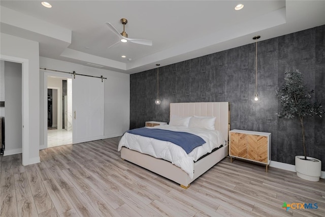 bedroom featuring a barn door, ceiling fan, a raised ceiling, and light hardwood / wood-style flooring