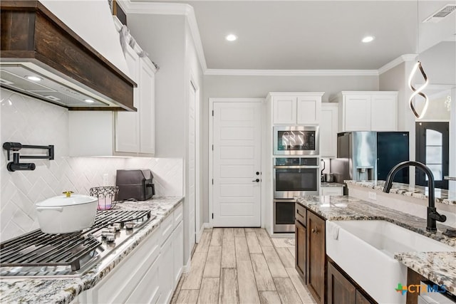 kitchen with appliances with stainless steel finishes, premium range hood, white cabinetry, ornamental molding, and tasteful backsplash
