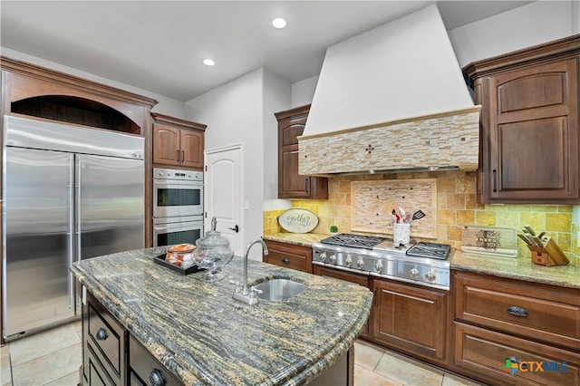 kitchen with premium range hood, sink, a center island with sink, dark stone countertops, and appliances with stainless steel finishes