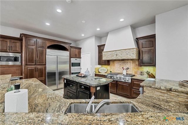 kitchen featuring premium range hood, sink, built in appliances, light stone countertops, and backsplash