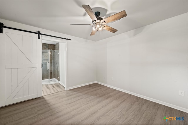 unfurnished bedroom featuring wood finished floors, ensuite bathroom, a barn door, baseboards, and ceiling fan
