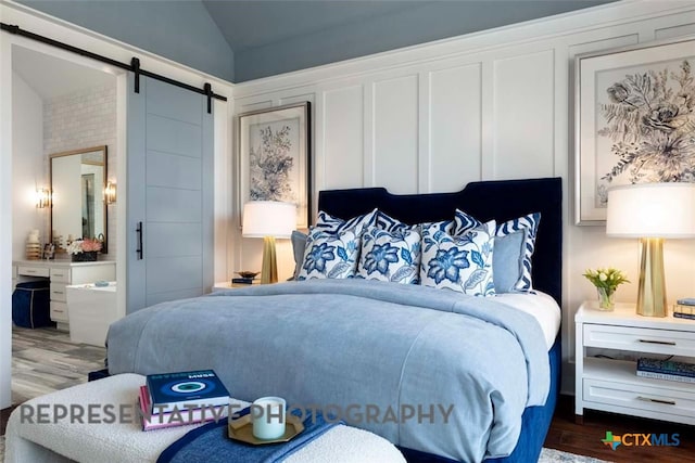 bedroom featuring wood finished floors, vaulted ceiling, ensuite bathroom, a barn door, and a decorative wall