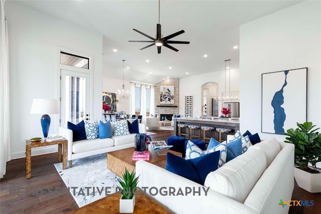 living area featuring a stone fireplace, recessed lighting, ceiling fan with notable chandelier, arched walkways, and dark wood-style floors