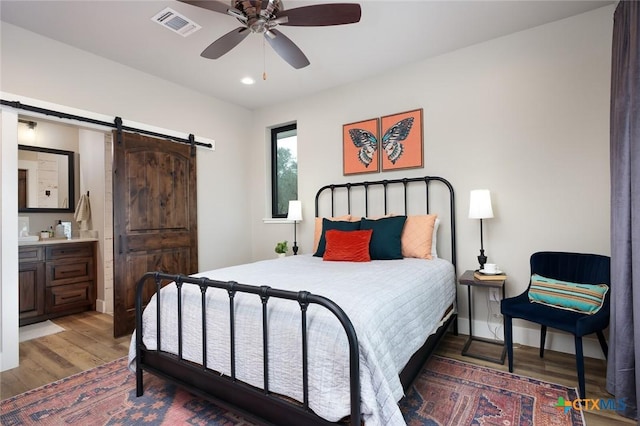 bedroom with ensuite bathroom, a barn door, dark wood-type flooring, and ceiling fan