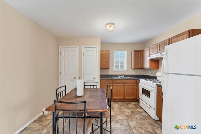 kitchen featuring white appliances, brown cabinetry, a sink, under cabinet range hood, and dark countertops