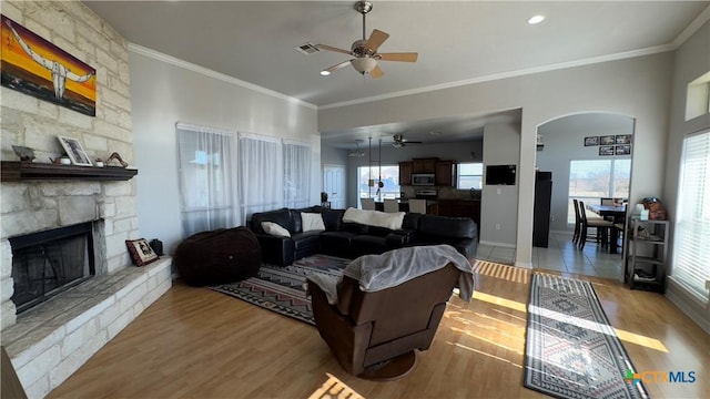 living room with a stone fireplace, ceiling fan, crown molding, and light wood-type flooring