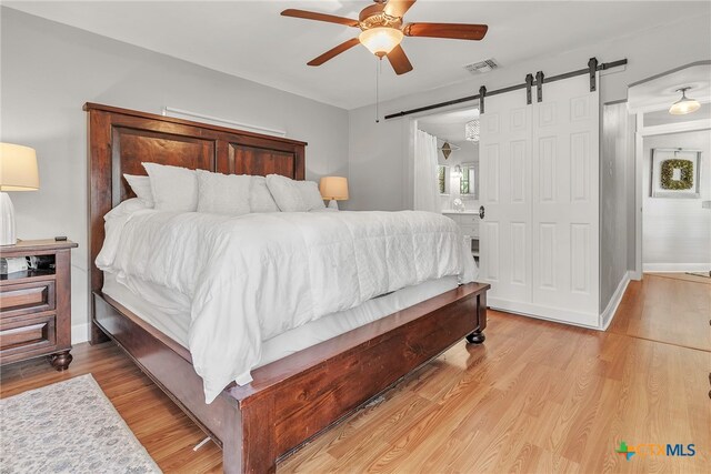 bedroom with connected bathroom, a barn door, ceiling fan, and light hardwood / wood-style flooring