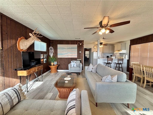 living room featuring ceiling fan, light wood-type flooring, and wooden walls