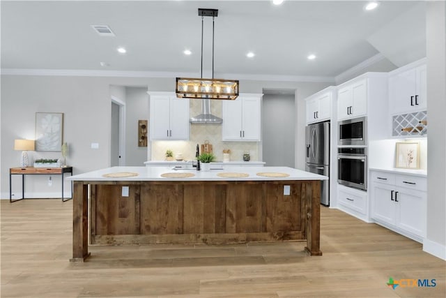 kitchen with pendant lighting, appliances with stainless steel finishes, a kitchen island with sink, white cabinets, and wall chimney exhaust hood