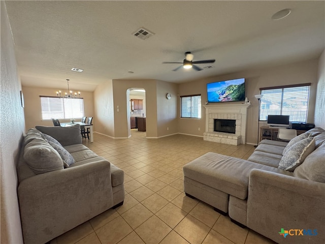 living room featuring ceiling fan with notable chandelier, a textured ceiling, a stone fireplace, and light tile patterned flooring