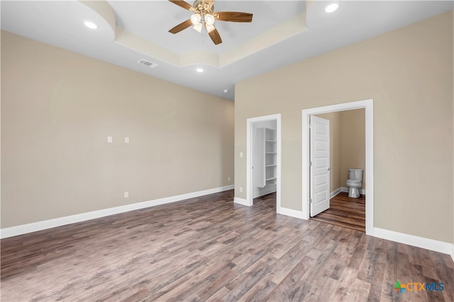 unfurnished bedroom featuring a spacious closet, ensuite bath, hardwood / wood-style floors, a tray ceiling, and ceiling fan