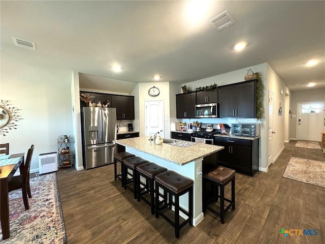 kitchen with a kitchen bar, appliances with stainless steel finishes, light stone counters, a center island with sink, and dark hardwood / wood-style floors