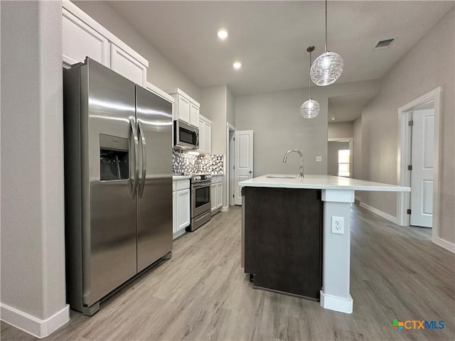 kitchen with a kitchen island with sink, stainless steel appliances, decorative backsplash, white cabinets, and decorative light fixtures