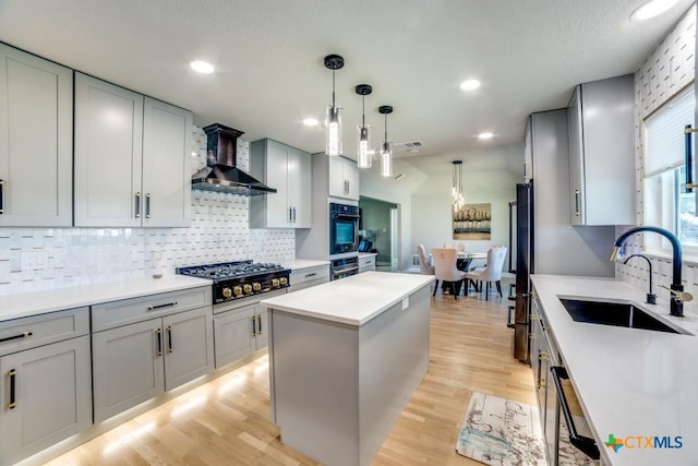 kitchen featuring light wood finished floors, a kitchen island, wall chimney range hood, black appliances, and a sink