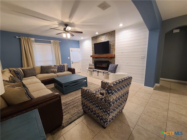 living room with a stone fireplace, ceiling fan, wooden walls, and light tile patterned floors