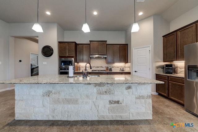 kitchen featuring tasteful backsplash, light stone counters, stainless steel appliances, decorative light fixtures, and an island with sink