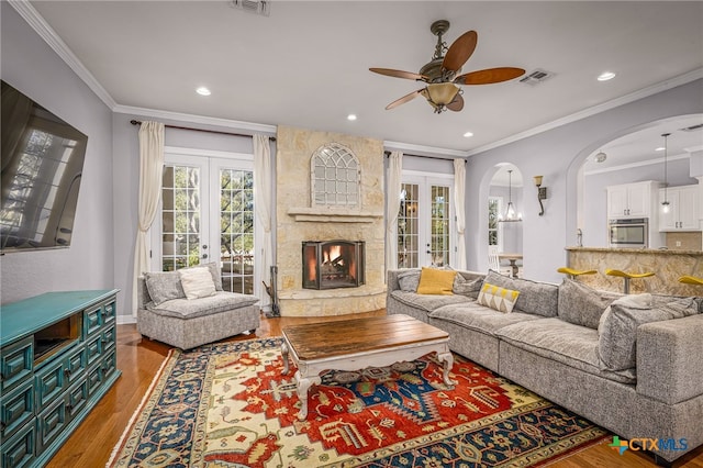 living room with french doors, a stone fireplace, ornamental molding, hardwood / wood-style flooring, and ceiling fan