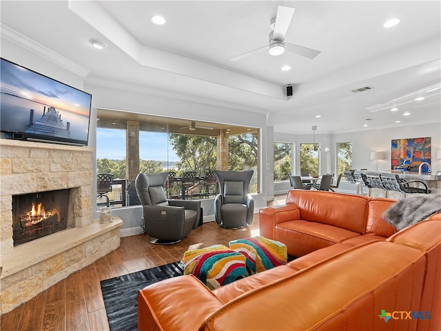 living room with a tray ceiling, a stone fireplace, ceiling fan, and crown molding