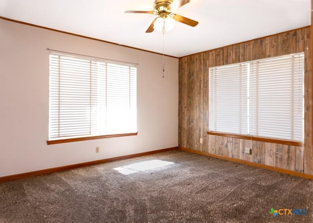 carpeted empty room with a ceiling fan, wood walls, baseboards, and ornamental molding