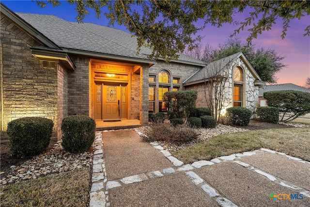 exterior entry at dusk featuring brick siding and a shingled roof