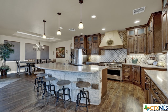 kitchen with dark hardwood / wood-style flooring, high end appliances, hanging light fixtures, custom exhaust hood, and an island with sink