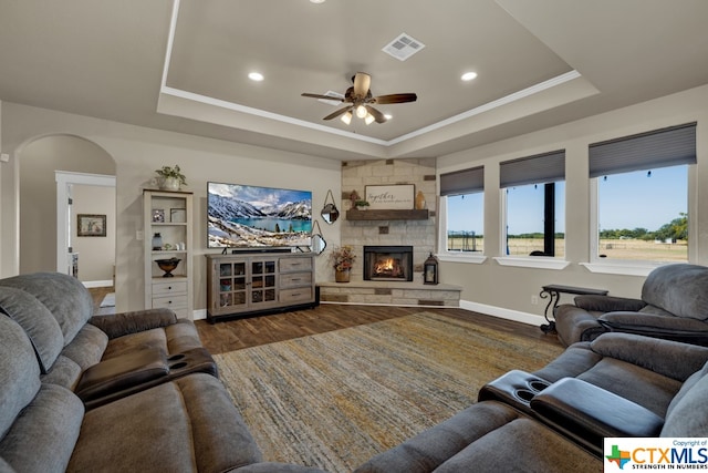 living room featuring a stone fireplace, ornamental molding, dark hardwood / wood-style floors, ceiling fan, and a tray ceiling