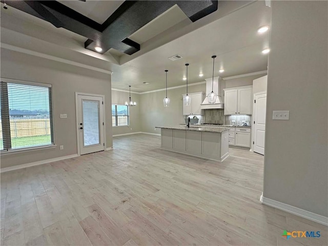 kitchen featuring light stone counters, hanging light fixtures, a center island with sink, custom range hood, and white cabinets