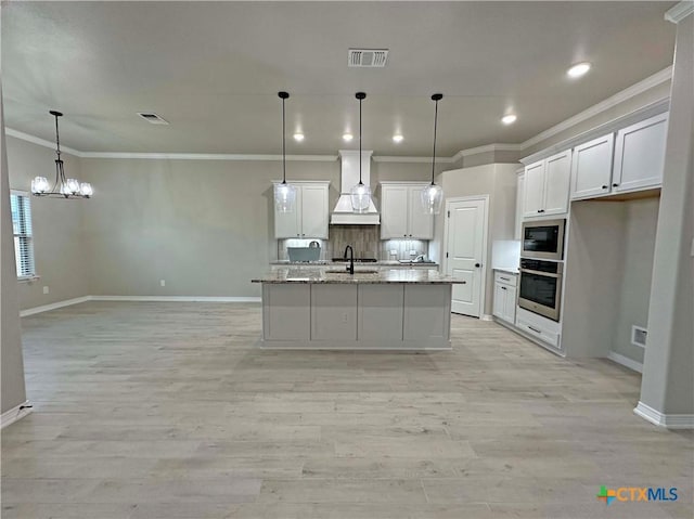 kitchen with premium range hood, pendant lighting, white cabinets, oven, and a kitchen island with sink