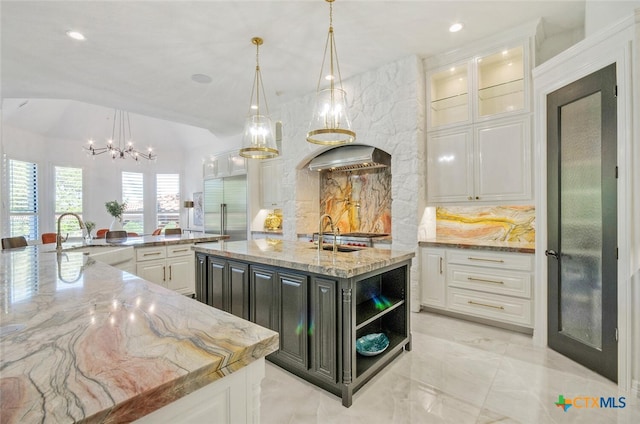 kitchen featuring light stone counters, built in refrigerator, hanging light fixtures, sink, and a large island with sink