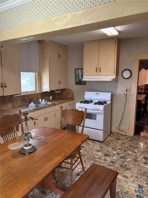 kitchen with under cabinet range hood, a sink, cream cabinetry, white gas range, and wallpapered walls