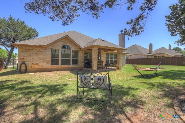 rear view of property with a shingled roof, a yard, stone siding, a chimney, and fence